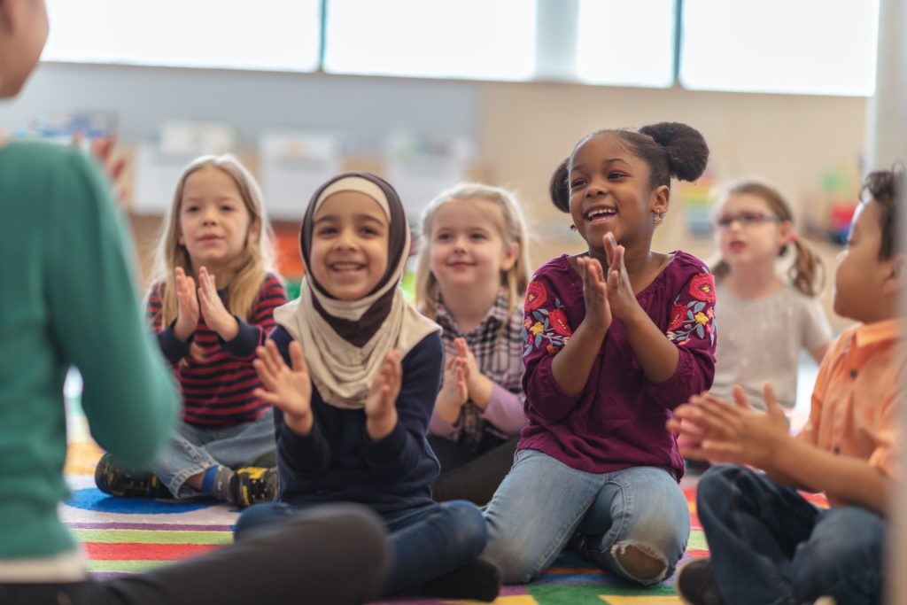 A group of kids are clapping along to music. They are sitting in their classroom.