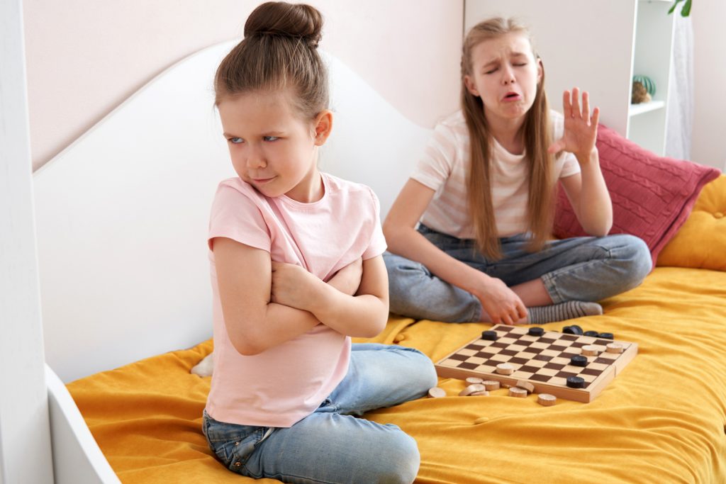 Two sisters playing checkers on a bad and having small fight How to Deal with a Sore Loser - SavvyMom