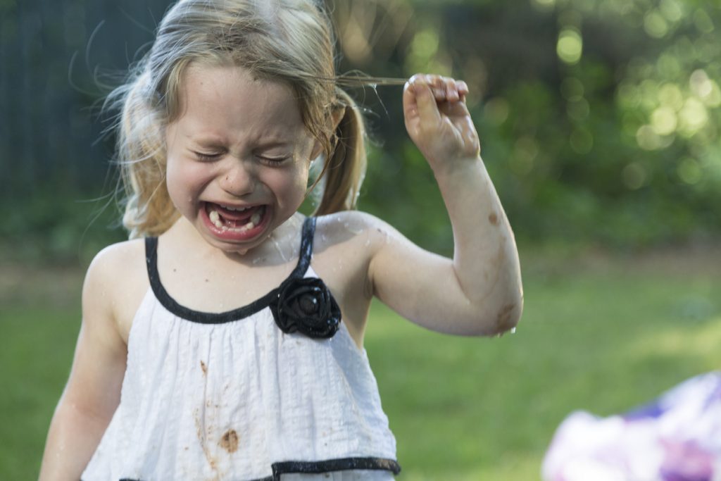 A little blonde girl with blue eyes and caucasian skin crying.  She is outside in the backyard and is very upset.  She has pig tails and she is a little muddy and damp.  She is wearing a white dress with black accents and is pulling her hair.  The background is green grass and trees.