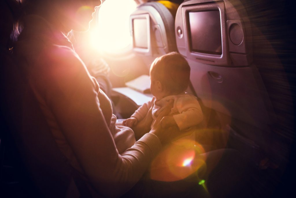 A mother and her baby girl sit in a passenger airline seat, the sun shining brightly in through the plane window.  While air travel with children can be difficult, both mom and child are content, the mother with a smile on her face. Horizontal image.  INTENTIONAL LENS FLARE.