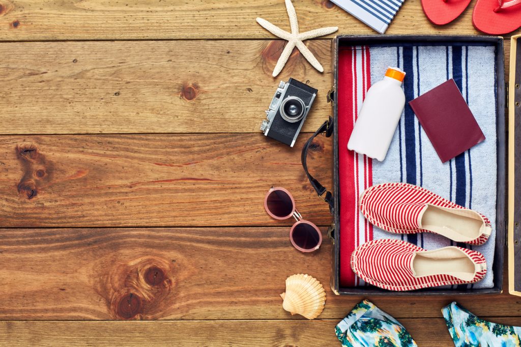 Directly above shot of open suitcase with summer vacation accessories. Flat lay of travel and beach equipment. They are placed on right side of hardwood floor. The copy space on floor can be used for advertisement purpose.