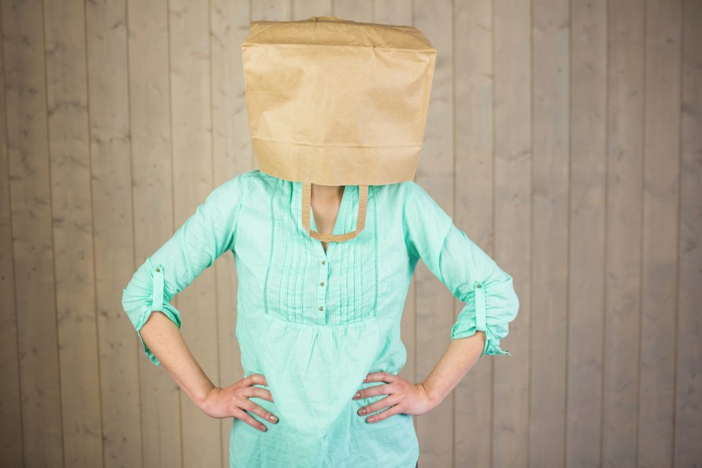 Woman covering head with brown paper bag while standing against wall