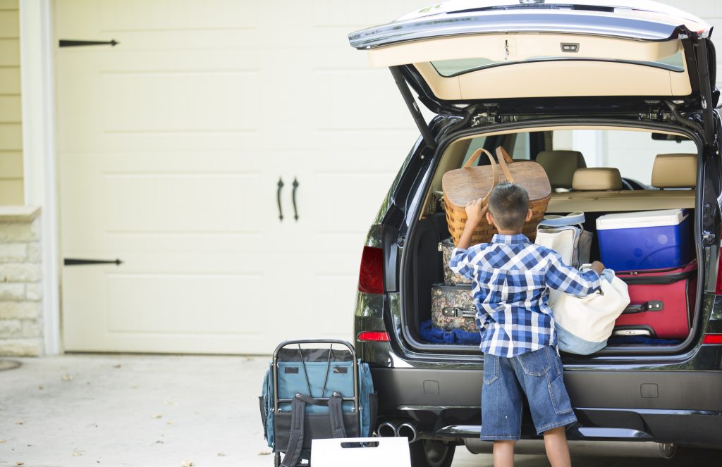 Family packs their vehicle for summer vacation.  One excited little boy waits by the car.