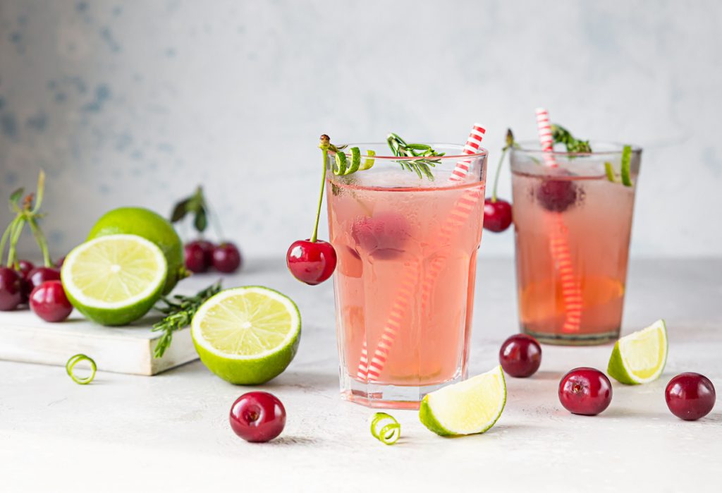 Cocktail or lemonade with cherries, lime and rosemary on a grey concrete table. Summer refreshment drink. Summer Cocktails - Sour Cherry Gin Smash - SavvyMom