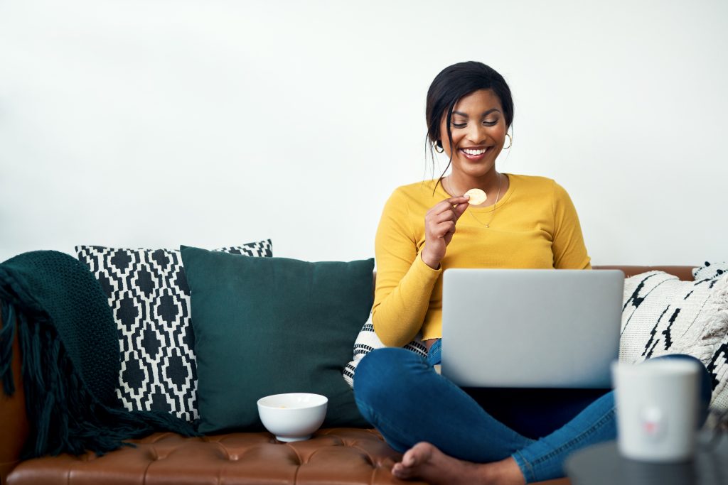 Cropped shot of an attractive young woman sitting on her sofa and eating potato crisps while using her laptop