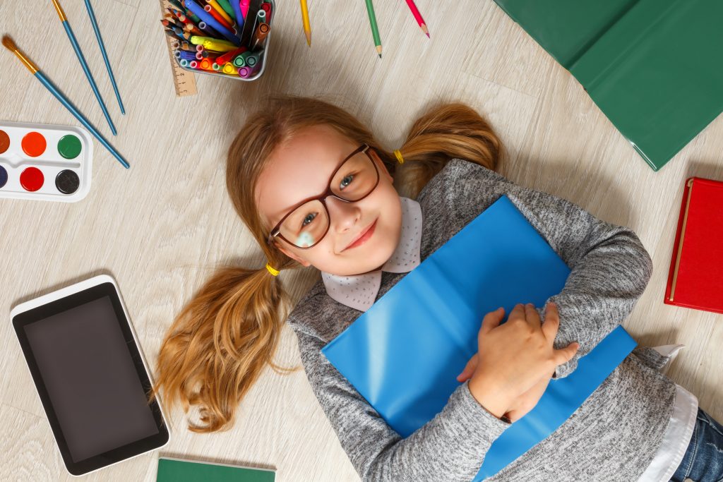 Cute little girl in glasses with a book lying on the floor. A child is surrounded by a book, tablet, paints, brushes, pencils. Cute little girl in glasses with a book lying on the floor. A child is surrounded by a book, tablet, paints, brushes, pencils.