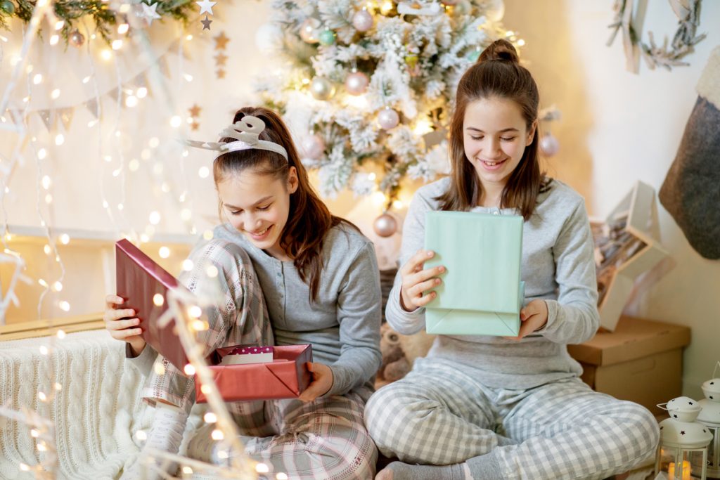 Two happy teenage girls in pajamas sitting by the Christmas tree and looking inside gift boxes
