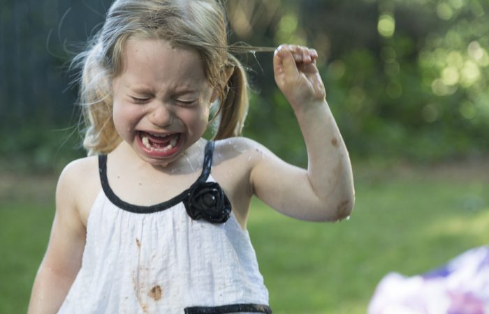 A little blonde girl with blue eyes and caucasian skin crying.  She is outside in the backyard and is very upset.  She has pig tails and she is a little muddy and damp.  She is wearing a white dress with black accents and is pulling her hair.  The background is green grass and trees.