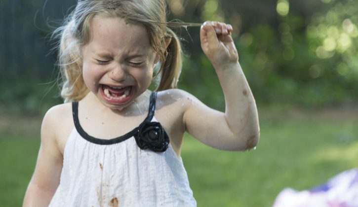 A little blonde girl with blue eyes and caucasian skin crying.  She is outside in the backyard and is very upset.  She has pig tails and she is a little muddy and damp.  She is wearing a white dress with black accents and is pulling her hair.  The background is green grass and trees.