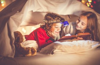 Two children are reading a book in bed under a sheet.  Little boy has a small headlamp on his head.  They are hiding under a homemade playing tent, made of bedding. Next to him, the little girl is holding her finger on the page of the book.