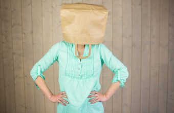 Woman covering head with brown paper bag while standing against wall