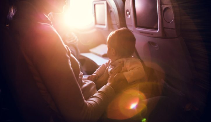 A mother and her baby girl sit in a passenger airline seat, the sun shining brightly in through the plane window.  While air travel with children can be difficult, both mom and child are content, the mother with a smile on her face. Horizontal image.  INTENTIONAL LENS FLARE.