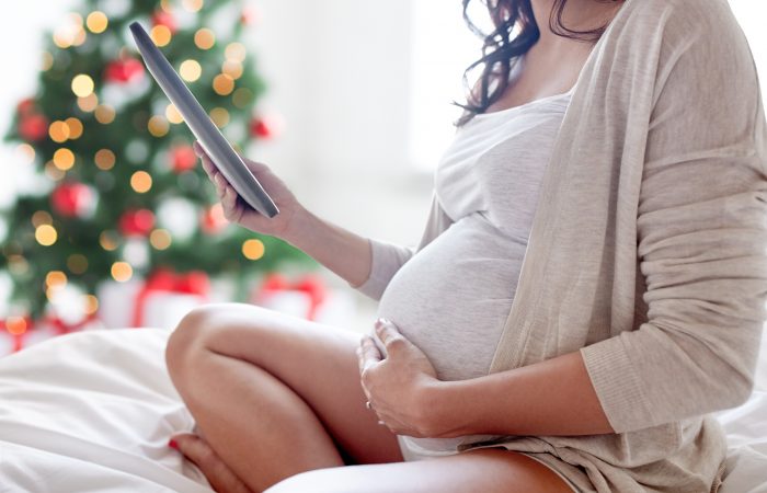 pregnancy, holidays, technology, people and expectation concept - close up of pregnant woman with tablet pc computer in bed at home over christmas tree background
