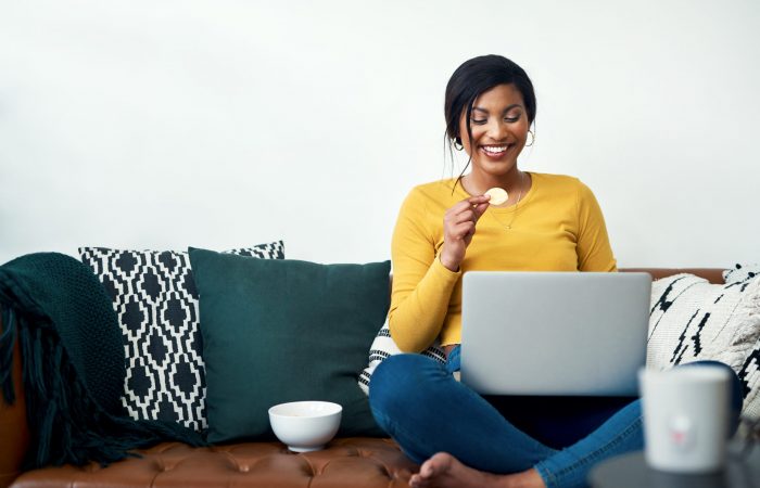 Cropped shot of an attractive young woman sitting on her sofa and eating potato crisps while using her laptop