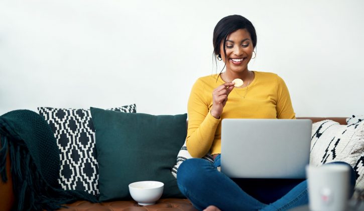 Cropped shot of an attractive young woman sitting on her sofa and eating potato crisps while using her laptop