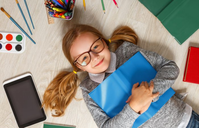 Cute little girl in glasses with a book lying on the floor. A child is surrounded by a book, tablet, paints, brushes, pencils.