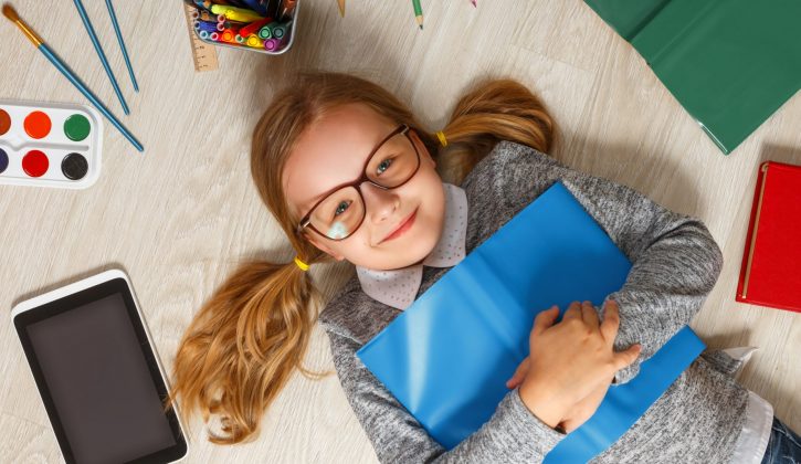 Cute little girl in glasses with a book lying on the floor. A child is surrounded by a book, tablet, paints, brushes, pencils.