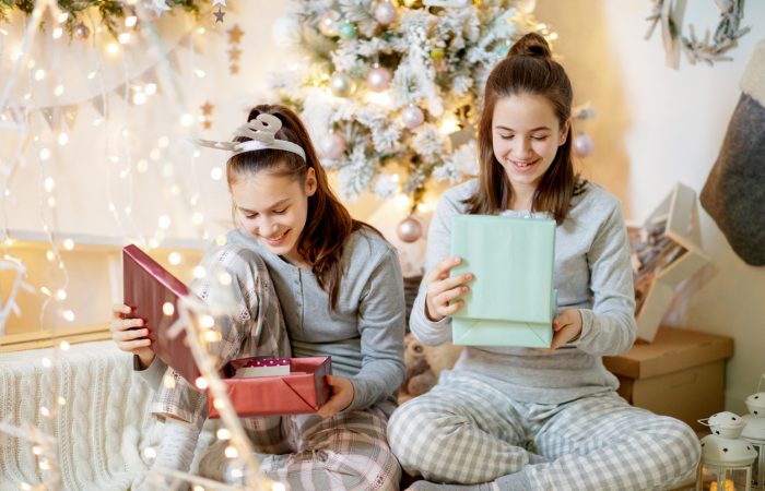Two happy teenage girls in pajamas sitting by the Christmas tree and looking inside gift boxes