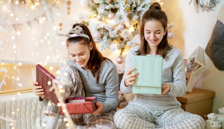 Two happy teenage girls in pajamas sitting by the Christmas tree and looking inside gift boxes